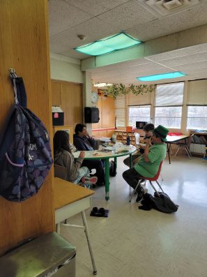 A group of people gathered around a table in a community space, engaged in conversation and activities, with bright windows and a warm atmosphere.